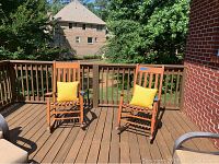 Two wooden rocking chairs with yellow cushions on an outdoor deck with wooden railing and greenery in the background.