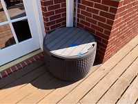 Photo of round rattan grill storage bin with grey wood plank patterned lid, placed outside on wooden deck near brick wall and glass door.