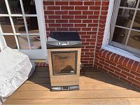 Front view of Masterbuilt smoker showing glass door and control panel on top, placed on wooden deck near brick wall.