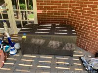 Rattan style pool storage box positioned outdoors on a patio with brick wall background. Shows dark brown woven texture and weathered condition.