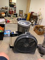 Photo shows Ninestar electronic trash can, Oreck Cyclone box fan with remote, and Lasko pedestal fan arranged on basement floor with other unrelated household items in background.
