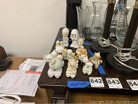 Front and slightly elevated view of 15 Precious Moments figurines arranged on a dark wooden surface, including bride and groom, baby figures, and bears on wheeled platforms