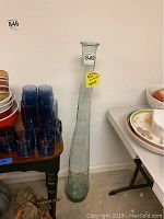 Tall narrow glass vase shown standing between a small table with blue glasses and a table with stacked plates, clearly showing its height and proportional width