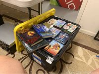 Photo of plastic storage bin with yellow lid, showing DVDs and books on top including Stephen King and Four Blind Mice books, various DVD cases underneath