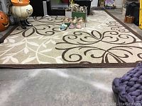 Full view of the 6x10 beige and brown leaf patterned polypropylene area rug displayed flat on concrete floor with some decorative figurines placed near the center.