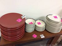 Red dinner plates stacked to the left, white bowls with silver rims in the center, and white heart-shaped plates stacked to the right on a wooden surface.
