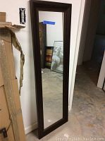 Two wall mirrors leaning against a white wall in a basement environment, one larger and one smaller, both with dark brown wooden frames.