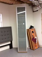 Front view of the tall shoe storage cabinet showing frosted glass door and wood grain sides, next to a cornhole game board and a bed frame.
