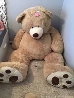 Full front view of the very large plush teddy bear seated on carpeted floor next to a drawer with water bottle in front for size comparison.