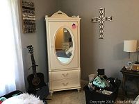 Front view of white Stanley armoire with oval mirror and two drawers, flanked by a black acoustic guitar.