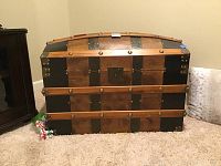 Front view of large wooden dome-top trunk with alternating wood and black metal panels, brass studs, metal reinforced corners, and central lock plate.