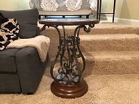 Front-side view of round table beside a sofa on a carpeted stair landing. Shows wooden top rim and metal base.