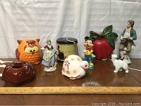 Photo showing assorted vintage knick knacks including ceramic owl, vintage lady figurine, cylindrical container, Mickey Mouse figurine, red apple container, seated male figurine, white cat figurine, floral turtle figurine, and brown glazed yarn holder.
