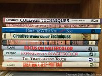 Photo of 10 watercolor and art technique books stacked on a wooden shelf with visible titles on spines.