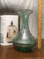Side view of the green hand-blown glass vase with a handpainted figure of a woman and a book in the background for scale.
