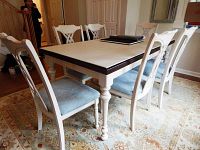 Six white wooden dining chairs placed around a matching dining table with a white and brown tabletop. Chairs have aqua chenille upholstered seats and elegantly turned legs.
