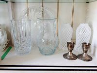 Display of items on shelf: sterling silver candlesticks, crystal cut vase, art glass vase, two milk glass hurricane shades, clear round serving platter