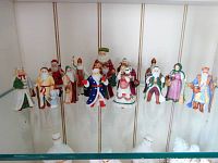 Photo showing 15 miniature porcelain Santa figurines arranged standing on a glass shelf, colorful and detailed uniforms, different poses including one with musical instruments and one female Santa.