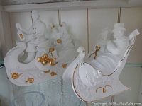 Front view of two white bisque Lenox musical Christmas figurines with couples in sleighs, detailed with gold accents and scrollwork, displayed on glass shelf.