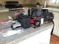 Overview of Sony camcorder, black case, cables, Canon DSLR body on box, two lenses, Tamrac bag on kitchen countertop