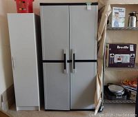 Front view of closed tall plastic storage cabinet with gray doors and black details next to a white cabinet and metal shelf unit.