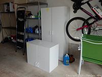 Two white laminate storage cabinets placed in a garage. Tall cabinet with double doors and four interior shelves; shorter cabinet also with double doors and two shelves. Cabinets shown next to other household items and a bicycle on the wall.