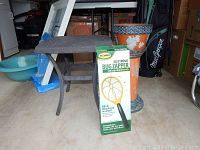 Photo showing a small square metal table, clay pedestal with pot and saucer behind it, and an electronic bug zapper box in front.