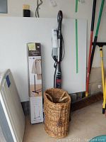 Photo showing the Shark Genius Steam Mop leaning against the wall with attached owner's guide, boxed portable garment rack, and woven wicker stair basket in front.