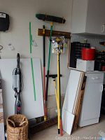 Photo showing outdoor brooms, large rake, pole lightbulb changer, and Euroshine squeegee hung on wall rack in utility area.