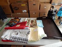 Wooden dark brown tray filled with assorted table linens in various colors and patterns on a white table with cardboard boxes in background