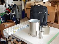 Photo showing handbags, golf clubs, cardboard boxes in background, and the primary lot items placed on a table including the suitcase, two window shades, and bathroom accessories.