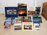 Photo showing a collection of assorted travel and history books including The Inventor Company, The Complete Pyramids, The Holy Land, History of Ireland, Lonely Planet Germany, Charleston, Amsterdam, Dartmoor, Santorini, and Winterthur Museum books.