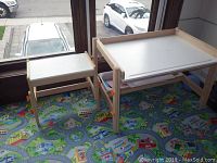 Two kids adjustable height tables, small and large, near a window on a colorful children’s play rug.