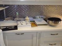 Overview of entire lot showing glass bowls, baking dishes, boxed espresso set, metal cheese platter, boxed salad servers, and glass vase on a kitchen counter