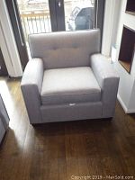 Front view of a light gray upholstered armchair with square arms and button-tufted back cushion sitting on hardwood floor