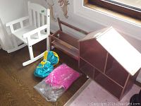 Photo showing white IKEA Sundvik children's rocking chair, small wooden bookshelf with two shelves, and a wooden dollhouse shaped like a house with multiple compartments, all placed on the floor near a window.