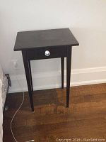 Pair of black wooden end tables with one drawer each, shown against a wall on hardwood floor. One table is missing a shelf and both show surface scratches.