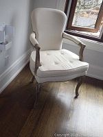 Front left angle view of white upholstered armchair showing tufted seat, wooden legs and armrests, and nailhead trim details.