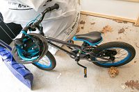 Bike folded sideways on the ground with visible blue tire rims, black frame marked Garneau F16, and a child-sized black and blue helmet hanging on the handlebar.