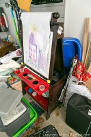 Full view of wooden children's easel with attached paper roll showing child's painting, surrounded by miscellaneous items in background.