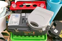 Closed black plastic tool box with handle and metal latches placed on green base with other tools nearby.