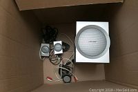 Three Bose speakers including one white subwoofer speaker with silver grille and two smaller silver satellite speakers, all placed inside a cardboard box, with cords coiled around them.