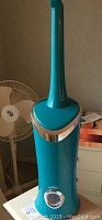 Full view of the tall teal humidifier with silver accent and dial control on base, placed in front of a white floor fan, showing its height and design.