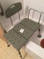 Grey bathtub seat with backrest and perforated seating surface inside a bathtub.
