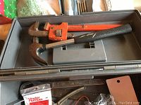 Open metal tool box showing hand tools including hammers, orange pipe wrench, and black handled hammer inside the upper compartment