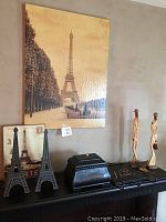 Image showing the group of items on a black mantel against a wall with Paris-themed artwork, two metal Eiffel tower candle holders, black leather box, and two tall wooden figurines.