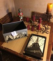 Decorative Paris motif storage box with lid removed, showing black and white Eiffel Tower images and metal hardware, alongside assorted candle holders on wooden table.