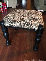 Front angle view of square foot stool with animal print fabric and black wooden legs on hardwood floor.