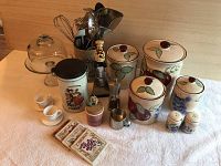 Four ceramic canisters with lids featuring fruit and floral designs, one has chip damage. Small ceramic salt and pepper shakers, two coasters, ceramic container labeled "The Best in Coffee," two small egg cups, and covered glass pedestal plate with clear dome cover are also displayed on a white cloth surface.