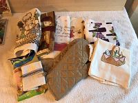 Photo shows multiple folded kitchen towels with designs, a beige quilted oven mitt, and packaged cutlery on a soft surface